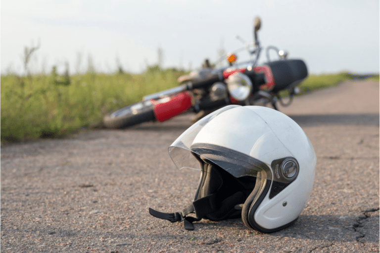 Close-up of a white helmet on the ground with a fallen motorcycle in the background on a rural road.